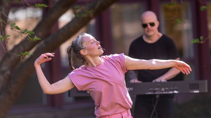 Kvinna med rosa tröja dansar utomhus. I bakgrunden syns en man med  solglasögon som spelar keyboard. 