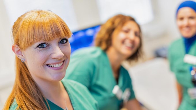 En rödhårig kvinna i gröna vårdkläder tittar in i kameran och ler. I bakgrunden syns två kvinnor som ler och som också är klädda i gröna vårdkläder.