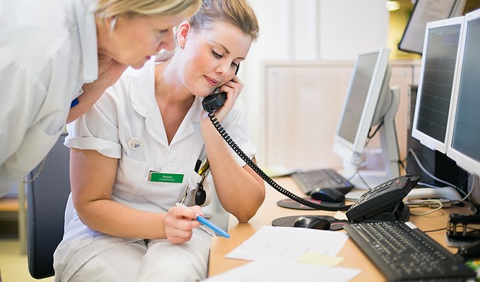 En sjuksköterska sitter och pratar i telefon vid ett skrivbord och läser på ett papper. En kollega lutar sig fram bredvid och läser på samma papper.