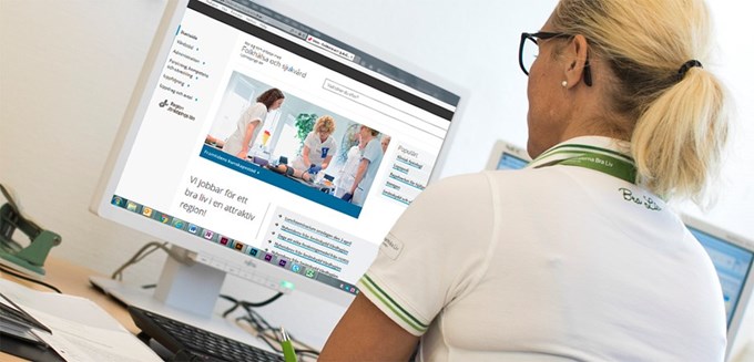En vårdklädd kvinnlig personal sitter framför en datorskärm. Datorskärmen visar den nya webbplatsen "Folkhälsa och sjukvård i Jönköpings län"