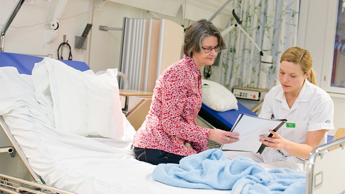 En kvinnlig patient sitter på sängkant i sjukhusmiljö. Hon tittar på ett dokument tillsammans med en kvinnlig vårdpersonal.