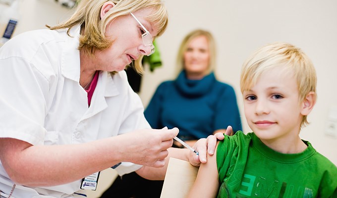 Barn får influensaspruta av kvinnlig sjuksköterska. I bakgrunden syns en annan vuxen, troligen barnets mamma
