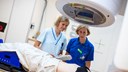 Två kvinnor iförda blå vårdkläder står vid en röntgenmaskin. De tittar ner på en person som ligger på en brits under maskinen.