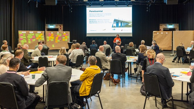 Personer som sitter runt bord i en stor sal och lyssnar till en talare, vid sidan om scenen står skärmar med post-it lappar. 