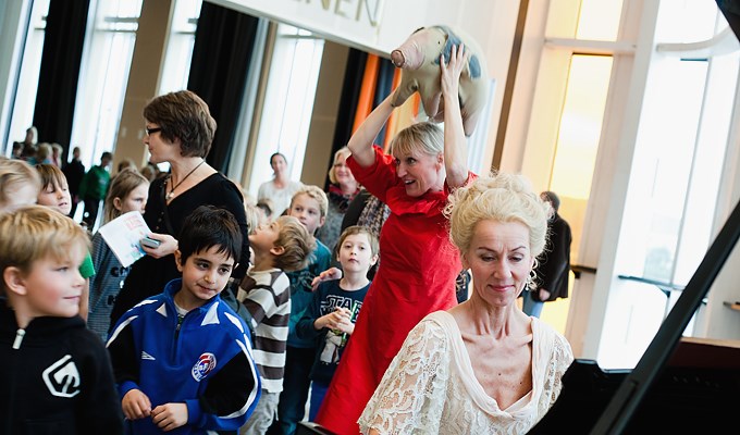 stor samling barn i kulturhuset spiras foajé tittar på en kvinna i röd klänning som håller upp en tyg-gris i luften. I förgrunden sitter en kvinna iklädd 1700-talskläder vid en flygel