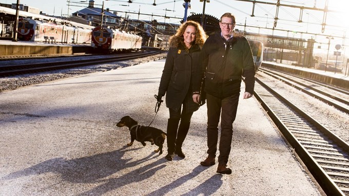 Man och kvinna med hund promenerar vid tågstation. 