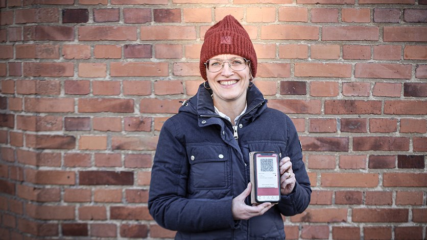 Porträtt på Helen Olofson. Hon står framför en tegelvägg och håller upp en smartphone med Jönköpings länstrafiks applikation. 