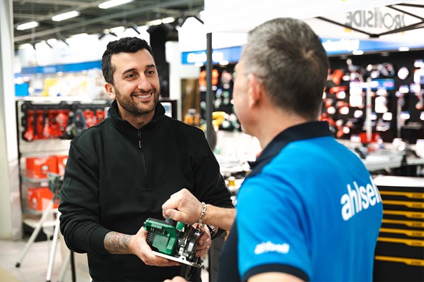 En man står i en byggvaruhandel och lämnar ett verktyg till butikspersonalen med ett stort leende.