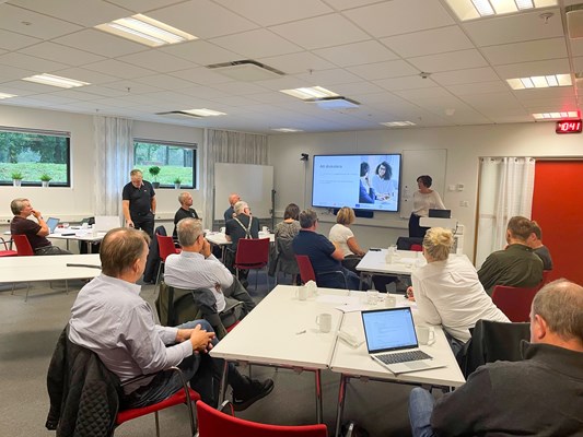 Personer sitter samlade runt bord och lyssnar