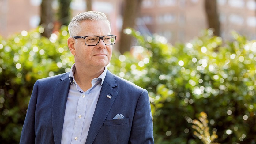 Peter står utomhus med grönt i bakgrunden och fint väder. Peter har en blå skjorta med en kavaj över.