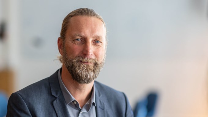 Harald som syns på bilden har uppsatt brunt hår och helskägg. Han är klädd i kavaj och ler mot kameran.