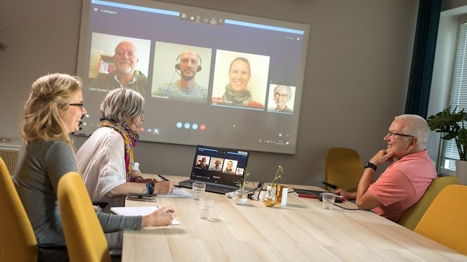 Resfritt digitalt möte med tre deltagare runt ett konferensbord och tre deltagare på en digital skärm.