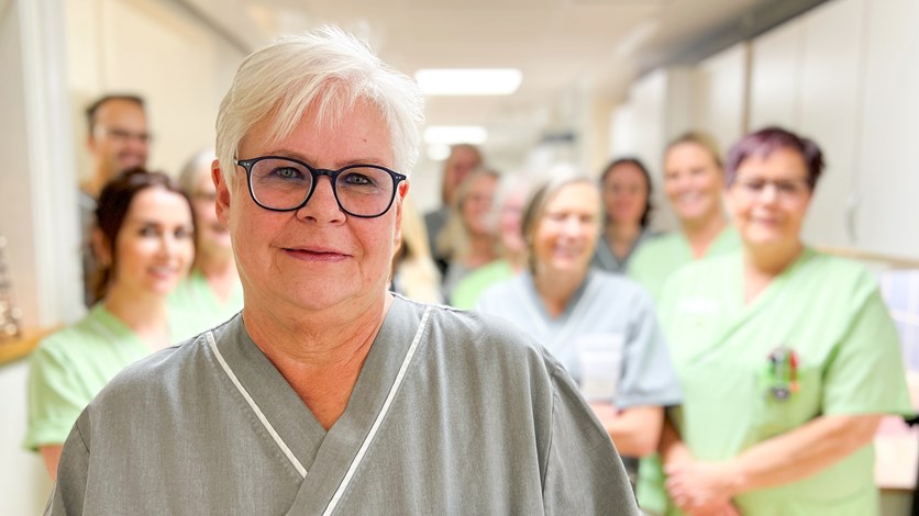 En kvinna står och ler in i kameran. Bakom henne syns ett gäng personer som också ler in i kameran.