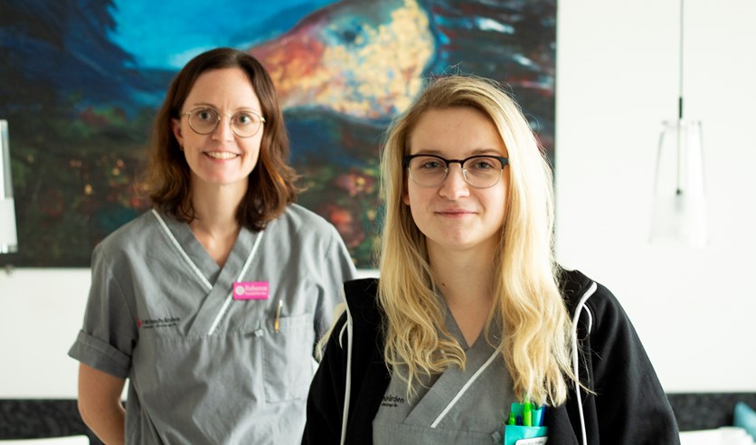 Två kvinnor står framför en stor målning och ler in i kameran. De har båda på sig en grå skjorta och glasögon. Kvinnan till vänster har brunt hår och kvinnan till höger är blond. 