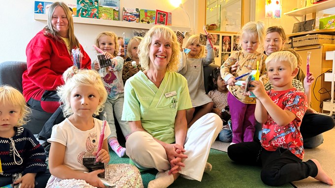 En kvinna sitter i mitten av ett glatt gäng med förskolebarn. Barnen håller i varsin tandborste, tandkräm och reflex.