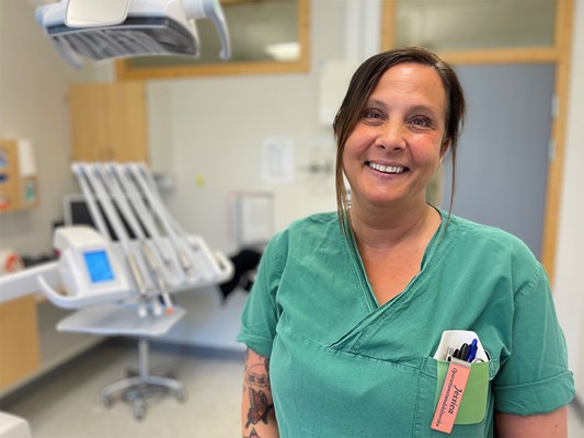 En kvinna med mörkt hår står i ett behandlingsrum iklädd klinikkläder.