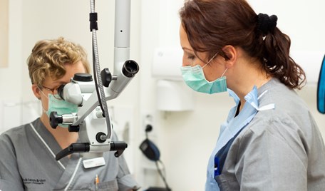 Två kvinnor med munskydd och grå tröja. Kvinnan till vänster tittar in i ett mikroskop.. 