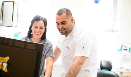 En kvinna och en man står lutade över en datorskärm. Båda är vårdklädda och står i ett kliniskt behandlingsrum.