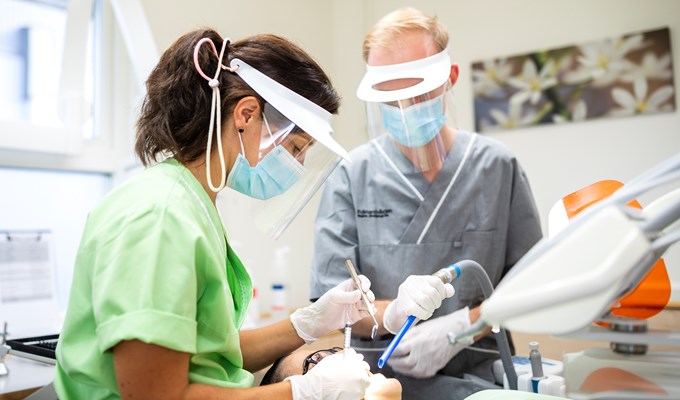 En kvinnlig tandläkare står böjd över en patient i behandlingsstolen. Bredvid henne står en manlig tandsköterska.