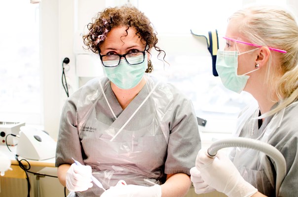 Bild på två kvinnor med munskydd och visir som utför en tandvårdsbehandling på en patient. 