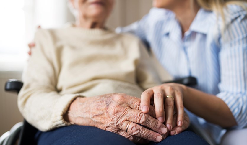 En ung kvinnas hand håller i en äldre kvinnas. Den äldre kvinnan sitter i rullstol och har båda händerna i knät. 