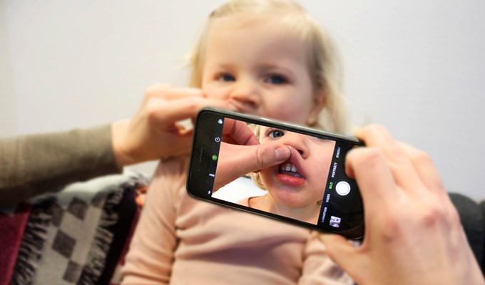 Ett barn sitter i en soffa. Framför henne håller någon i en mobiltelefon och tar ett foto med ena handen samtidigt som hon lyfter på läppen. 