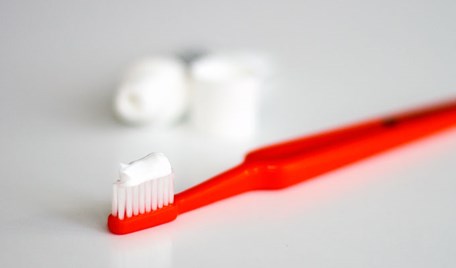 En röd tandborste ligger på ett bord. I bakgrunden syns en tandkräm.