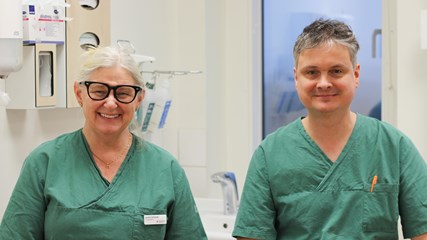 En man och en kvinna sitter i ett behandlingsrum. De har gröna klinikkläder på sig. De ler och tittar in i kameran.