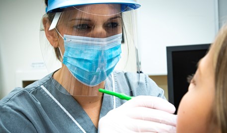 Närbild på kvinna med visir och munskydd som undersöker munnen på en patient. Bakhuvudet på patienten har brunt hår och syns i förgrunden.