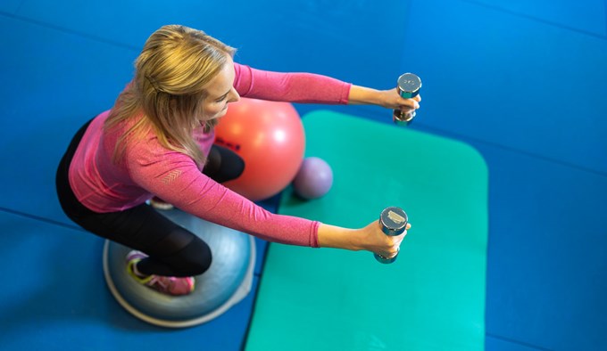 En kvinna med rosa långärmad träningströja som står på en balansboll framför en grön träningsmatta. 