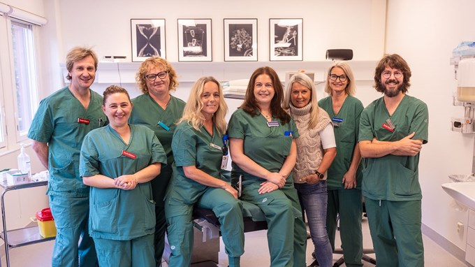 Åtta medarbetare på sjukhuset står bredvid varandra och ler mot kameran. De har vårdkläder på sig. 