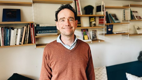 En person ler mot kameran. Personen står i ett bibliotek. 