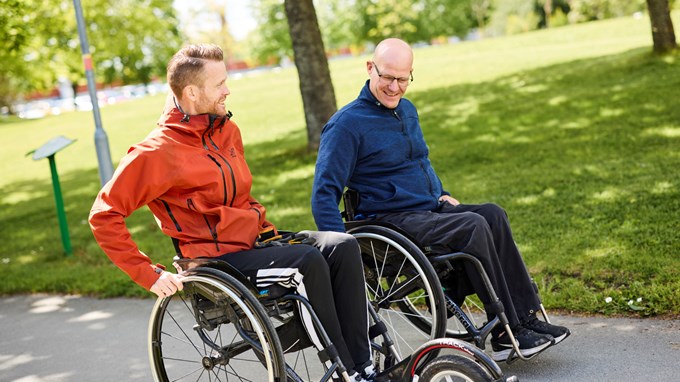 Två personer i rullstol pratar och rör sig längs en gångväg i en park.