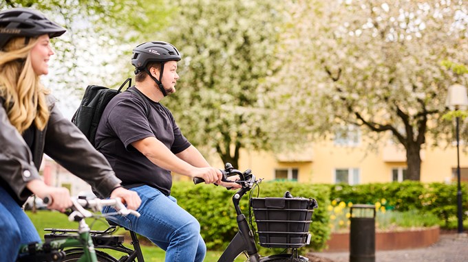 Två personer cyklar tillsammans på en cykelbana i stadsmiljö, båda bär cykelhjälm.