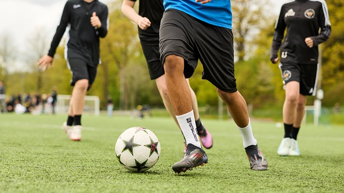 Flera personer spelar fotboll på en plan. En spelare i blå tröja driver bollen framåt.