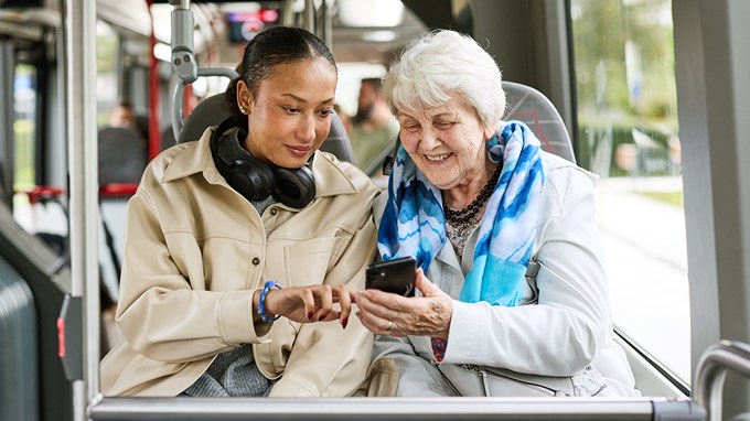 En yngre och en äldre kvinna sitter på en buss och tittar tillsammans på en mobiltelefon.