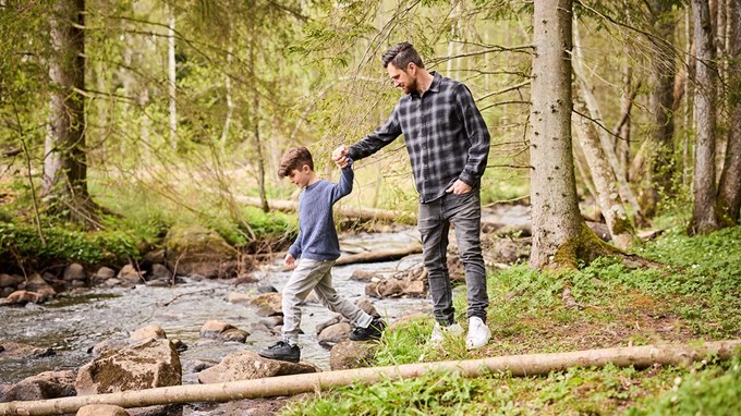 En vuxen och ett barn går över en bäck i en skog. Den vuxne håller barnet i handen för att hjälpa det balansera på stenarna. 