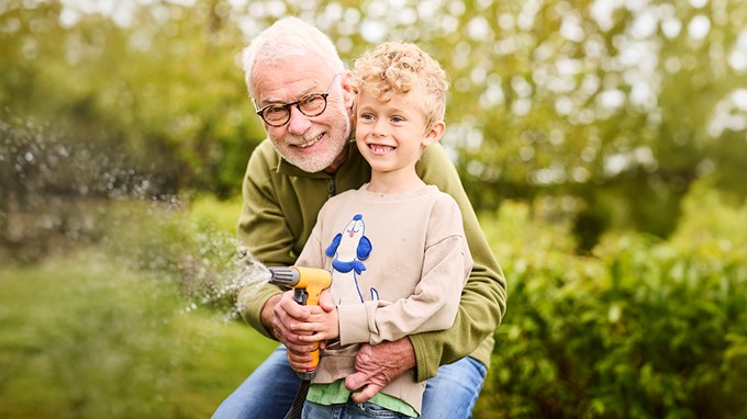 En äldre man och ett barn vattnar i trädgården tillsammans med en trädgårdsslang.