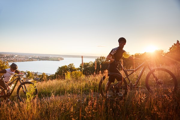 Två cyklister leder sina mountainbikes uppför en äng i tidigt morgonljus. I bakgrunden  syns Huskvarna och Jönköping vid Vätterns strand.