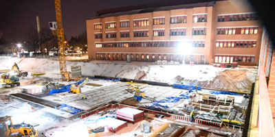 Upplyst byggarbetsplats, med lyftkran och grävmaskiner. I bakgrunden syns det befintliga Länssjukhuset Ryhov.