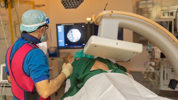 Person i vårdkläder och skyddsväst genomför en undersökning med röntgenapparat på en person som ligger ner.