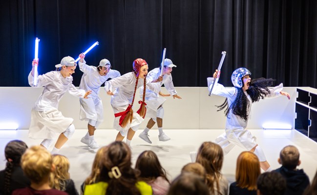 Fem vitklädda skådespelare som springer med stavar i händerna på en scen.