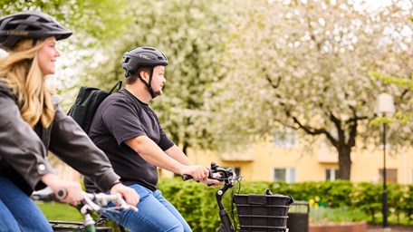 En man och en kvinna cyklar i ett bostadsområde. Grönska i bakgrunden. 