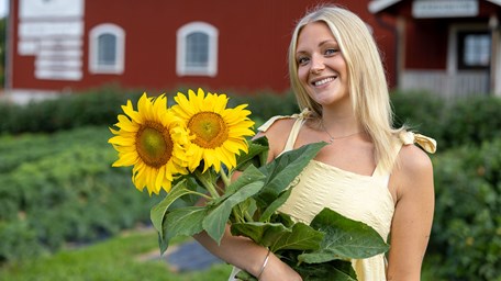 Ung kvinna med lång ljust hår håller i gula solrosor och ler mot kameran. 