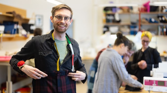 En ung man med glasögon ler in kameran. Han har ett måttband om halsen och visar upp et par byxor. 