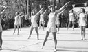 Gymnastikuppvisning på större träscen uppbyggd i Apladalen i Värnamo. Över scenen hänger vimplar. På bilden ses ett 10-tal flickor i gymnastikdräkt; scenen kantas av publik. Bild från 1940-talet. 