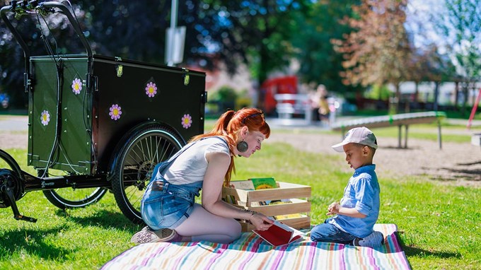 Bibliotekarie som sitter på en filt i en park och läser ur en bok för ett barn.