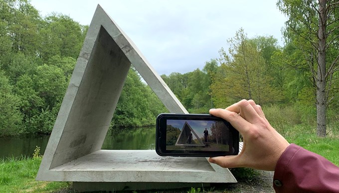 Ett konstverk i grå betong i form av en triangel. I förgrunden ses en hand som håller i en mobil för att fotografera konstverket.