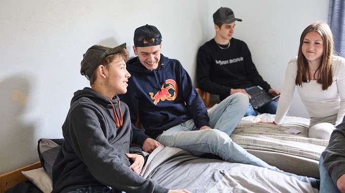 Två killar och en tjej sitter på en säng och skrattar. I bakgrunden syns en kille som läser på en dator.