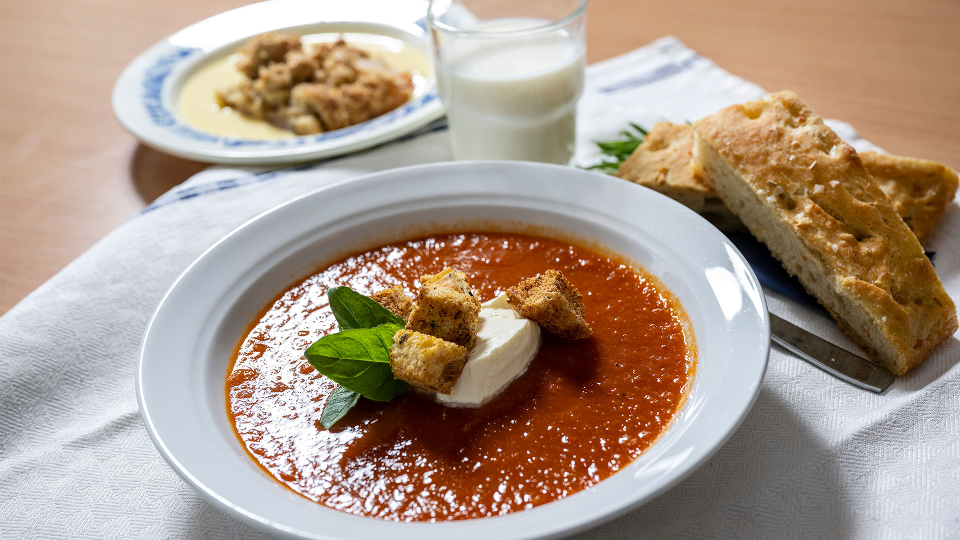 En tallrik med tomatsoppa och krutonger på, ett glas mjölk står bakom och lite bröd vid sidan om.